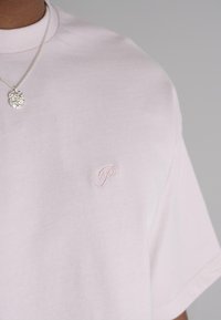 Light pink cotton t-shirt featuring a small, embroidered logo on the left chest. Loose fit with short sleeves and a ribbed crew neckline.