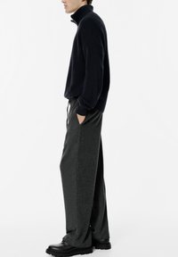 Sötét garbóban, szürke széles lábú nadrágban, amelyhez zsinór is tartozik, és fekete masszív cipőben álló személy, miközben a zsebébe dugja a kezét egy fehér háttér előtt.