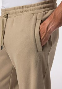 Khaki tepláky s elastick�ým pasem, bočními kapsami a malým embosovaným logem. Tkanina vypadá jemně a má hladký povrch.