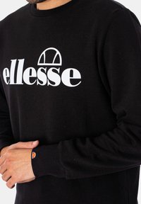 Schwarzer Sweatshirt mit einem herausragenden weißen "ellesse"-Logo. Die Bündchen haben einen farbigen Logo-Akzent. Das Material wirkt weich mit einer strukturierten Oberfläche.