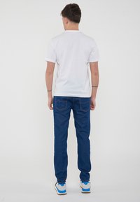 T-shirt bianco a maniche corte, jeans in denim blu con cuciture rosse sui taschini, indossati con scarpe da ginnastica grigie e blu, persona vista da dietro.