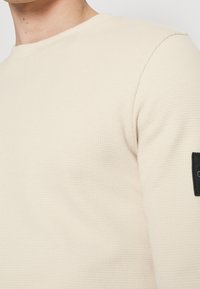 Beige rundhalströja gjord av texturerad tyg; har en svart patch på ärmen med varumärkeslogotyp.