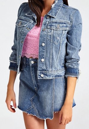 Kobieta nosząca niebieską jeansową kurtkę i asymetryczną jeansową spódnicę z różowym koronkowym krótki topem pod spodem.