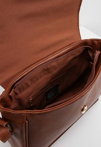 Ruskea nahkalaukku, jossa on sileä pinta, varustettuna vetoketjullisella sisätaskulla ja kultaisilla yksityiskohdilla. Aukileveä läppä paljastaa tilavan osaston.