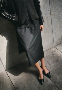 Falda negra de longitud media hecha de material suave, con una cintura texturizada. Combinada con zapatos negros puntiagudos, sobre un fondo de pared gris claro.