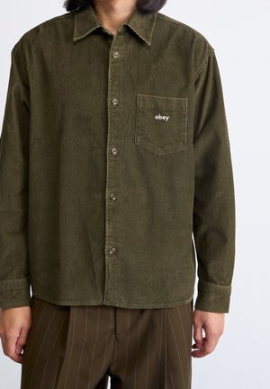 Person trägt ein olivgrünes Cordhemd mit Knöpfen und einer Brusttasche, bestickt mit "obey", kombiniert mit braunen Nadelstreifenhosen.