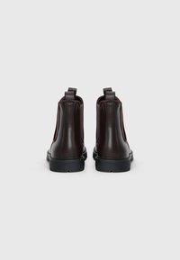 Botas de tobillo de cuero marrón con paneles laterales elásticos y tiradores. Suelas de goma negra y acabado texturizado. Vista trasera.