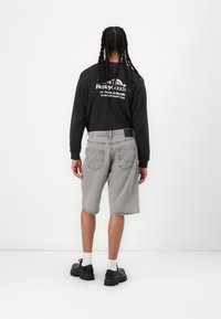 Musta pitkähihaisen paita, jossa on valkoinen logo, yhdistettynä harmaisiin denimpö shorts. Malli käyttää mustia kenkiä ja valkoisia sukkia, kääntyen pois valkoista taustaa vasten.