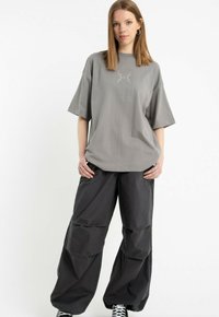 Młoda kobieta stoi w oversize'owej szarej koszulce z dekoracyjnym wzorem oraz luźnych czarnych spodniach cargo, w sneakersach, na białym tle.