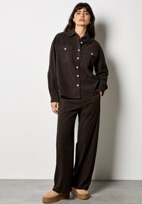 Camisa de pana marrón con botones y dos bolsillos en el pecho, combinada con pantalones de pernera ancha a juego y zapatos de plataforma beige, sobre un fondo neutro.