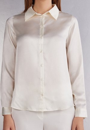 Woman wearing long-sleeve, cream-colored satin blouse with pointed collar and front buttons, paired with matching cream pants against plain background.