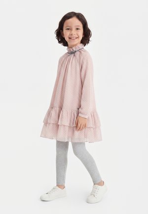 Hymyilevä nuori tyttö seisoo sivuttain, päällään vaaleanpunainen röyhelömekko, harmaat leggingsit ja valkoiset lenkkarit yksiväristä valkoista taustaa vasten.