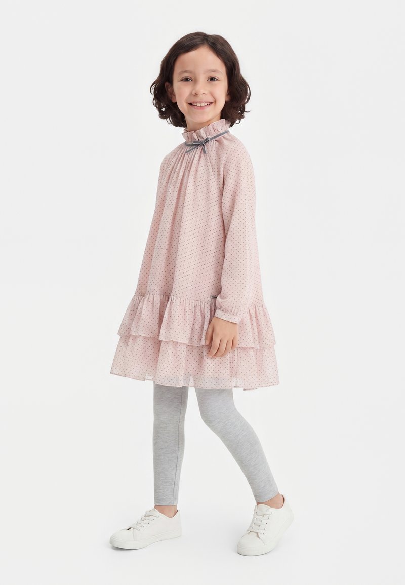 Jeune fille souriante debout de profil, vêtue d'une robe rose à volants, de leggings gris et de baskets blanches sur un fond blanc uni.