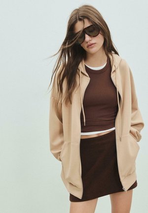 Mladá žena s dlhými vlasmi, oblečená v presvetlenom béžovom zip-up svetri, hnedom krátkom topu, zodpovedajúcej sukni a veľkých tmavých slnečných okuliaroch, stojí proti jednému pozadiu.