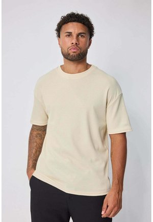 Homme aux cheveux bouclés et barbe, portant une chemise beige à manches courtes texturée et un pantalon noir, debout avec une main dans la poche.