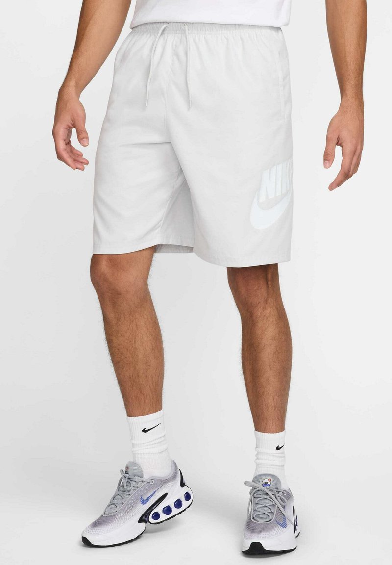 Άνδρας που φοράει λευκά σορτς Nike, λευκές κάλτσες Nike και γκρι παπούτσια τρεξίματος Nike, στέκεται μπροστά σε έναν απλό φόντο.