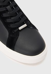 Zwarte sneaker met een soepel leren bovenwerk, suède accenten, zwarte veters en een witte rubberen zool met een slanke, ronde neusdesign.