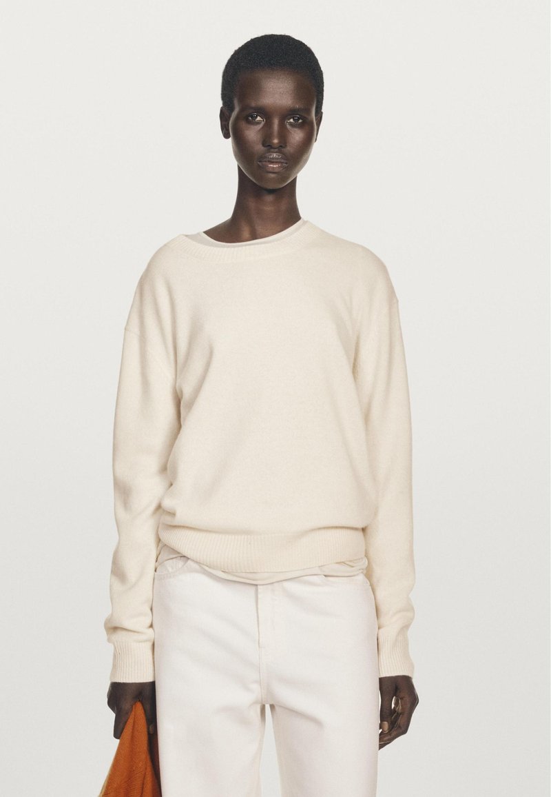 Light beige knit sweater with a round neckline and ribbed cuffs and hem, paired with white trousers, held with an orange accessory.
