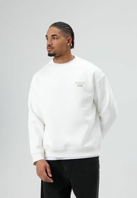 Witte sweat met een ronde hals en geribbelde manchetten. Heeft het geborduurde logo "dreams Chasing" in lichtgrijs. Zwarte broek, neutrale achtergrond.