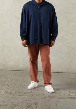 Mężczyzna w granatowej kurtce zapinanej na guziki, brązowych spodniach i białych trampkach, stojący na teksturowanej betonowej podłodze na tle neutralnej ściany.