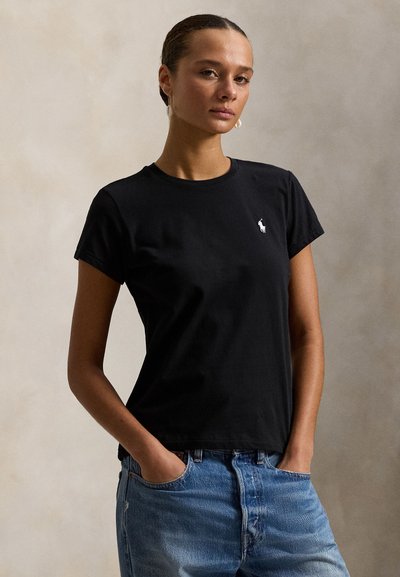 Femme aux cheveux tirés en arrière portant un t-shirt noir avec un petit logo blanc et un jean bleu, debout les mains dans les poches devant un fond uni.