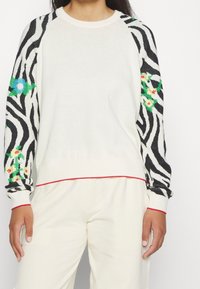 Sweatshirt blanc avec des manches tricotées à rayures zébrées noires ornées de motifs floraux colorés. Bordure rouge aux poignets et à la taille.