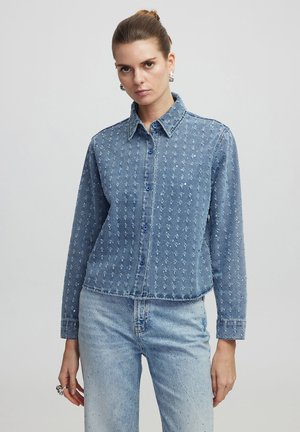 Camicia di jeans azzurro chiaro con dettagli ricamati, design con colletto, maniche lunghe, chiusura a bottoni e un fit leggermente accorciato.