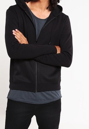 Haut à capuche zippé noir avec une poche avant et un cordon de serrage, porté sur un t-shirt gris à col rond. Tissu doux et lisse avec une coupe ajustée.