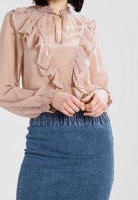 Blusa trasparente color pesca con dettagli in ruffle e collo a fiocco, abbinata a una gonna a tubino in denim scuro aderente con vita arricciata.