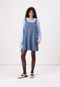 Denim A-line kjole med justerbare stropper, lagd over en blå og hvit stripete bluse. Modell med en hvit håndveske og svarte strømpebukser.