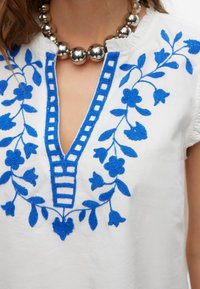Witte katoenen blouse met ingewikkelde blauwe bloemenborduurwerk langs de halslijn en zijkanten, met een diepe V-hals en een textuursteekdetail.
