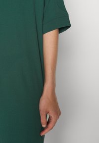 Bras droit et main d'une personne portant un vêtement à manches courtes vert foncé sur un fond clair uni.