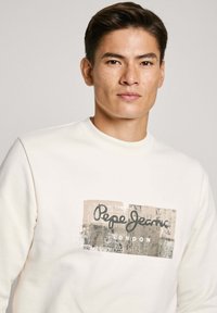 Sudadera blanca con una impresión gráfica que presenta "Pepe Jeans London" en texto negro sobre un fondo beige. Textura suave y diseño clásico de cuello redondo.