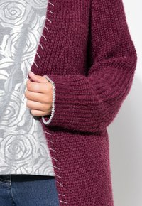 Bordový pletený cardigan s texturovaným designem, kontrastním prošíváním a volným střihem. V kombinaci se šedým topem s květinovým vzorem.