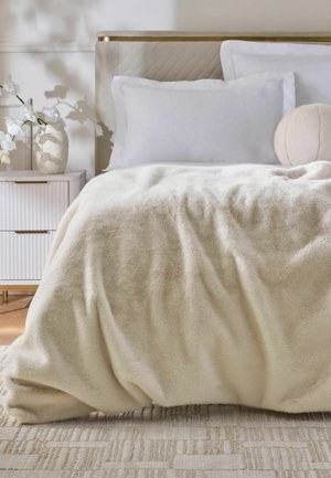 Coperta beige morbida sul letto con cuscini bianchi, cuscino rotondo, testiera beige, comodino bianco e orchidea bianca in un vaso.