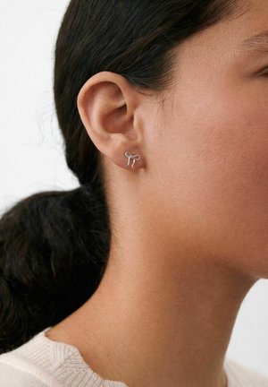 Kvinde med mørkt hår bundet tilbage, iført en lille sølv ørestik formet som en sløjfe på venstre øre.