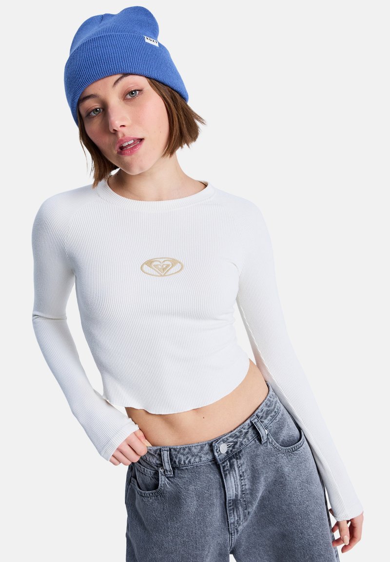Λευκό ριμπ μακρυμάνικο crop top με χρυσό λογότυπο, σε συνδυασμό με ψηλόμεσα γκρι τζιν. Το μοντέλο φοράει μπλε πλεκτό σκουφάκι.