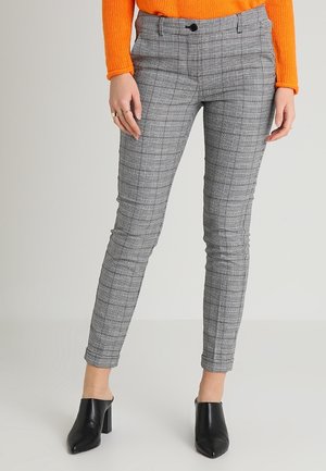 Nainen, joka pitää yllään kapeat harmaat ruudulliset housut, oranssin villapaidan ja mustat korkokengät, seisoo yksinkertaisen vaalean taustan edessä.