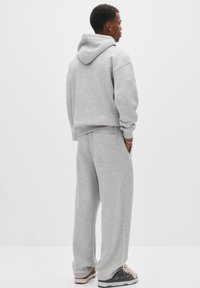 Ung mand står med siden til, iført en lysegrå hættetrøje og matchende sweatpants samt sorte og hvide sneakers på en hvid baggrund.