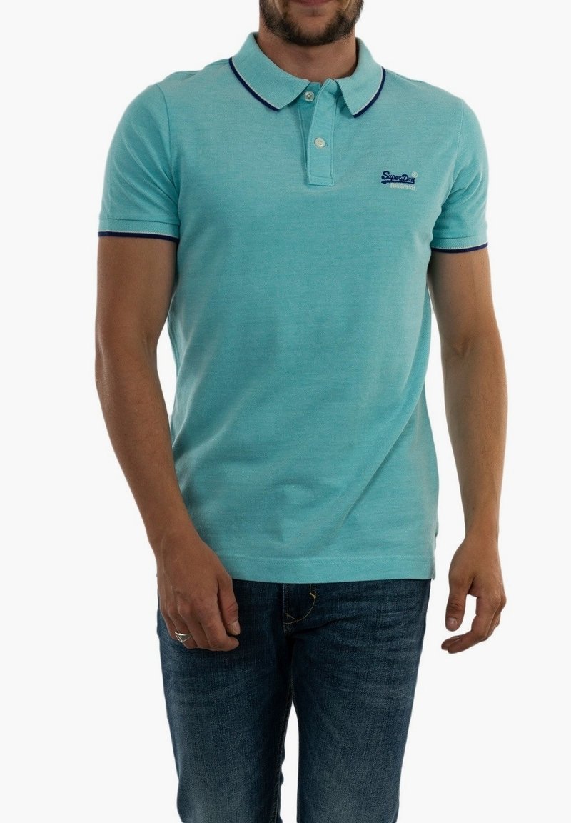 Homme portant un polo bleu clair avec des bordures foncées et un jean bleu, debout devant un fond blanc uni.