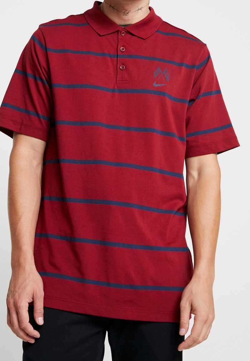 Polo rouge avec des rayures horizontales bleu marine, manches courtes, trois boutons et un petit logo sur la poitrine gauche. Matière en coton avec une texture lisse.