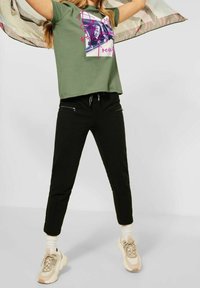 Grön bomull t-shirt med grafiskt tryck, tillsammans med svarta byxor med fickor med dragkedja och ljusa klumpiga sneakers. Halsduk i rörelse.