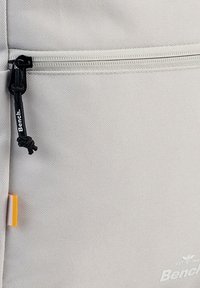 Sac à dos en tissu gris clair avec fermeture éclair supérieure, surface texturée et tirette de fermeture éclair noire portant le nom de la marque "Bench."