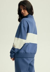 Niebieska bluza z wysokim kołnierzem i białym poziomym paskiem. Posiada ribbowe mankiety i dół, miękką tkaninę, luźny krój i minimalne detale.