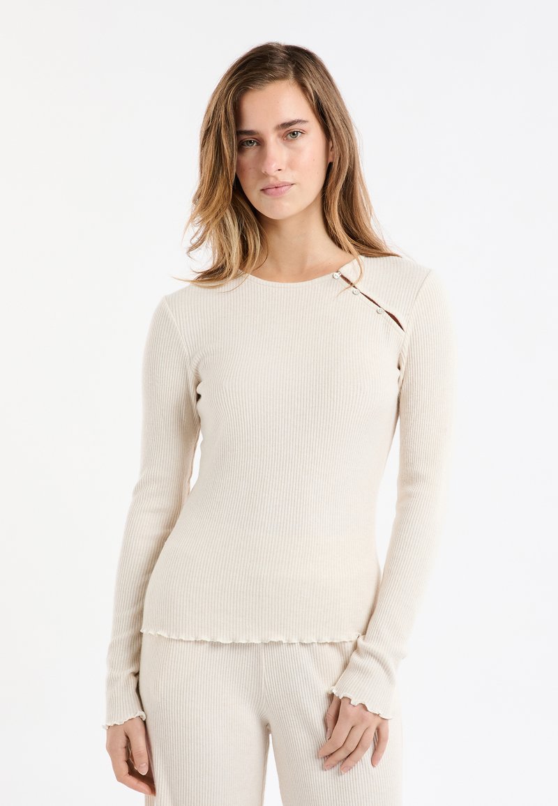 Top a maniche lunghe a coste beige con scollo rotondo, caratterizzato da bottoni decorativi sulla spalla e polsini arricciati. Tessuto morbido.