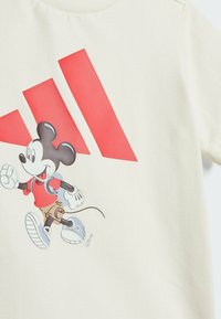 Camiseta de algodón color crema con un gráfico de un ratón de dibujos animados corriendo en rojo y beige, con zapatos azules, acentuada por rayas rojas bold.