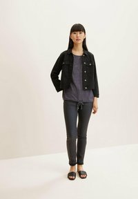 Jachetă din denim neagră peste un tricou purpuriu cu text, asociată cu blugi strâmți negri și sandale negre tip slide. Texturi netede și design ajustat.