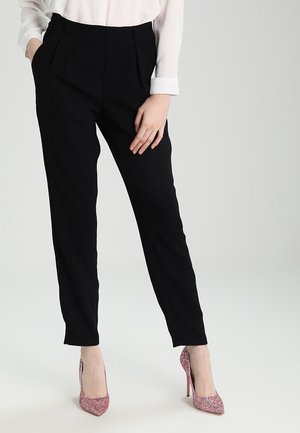 Žena stojící s jednou rukou v kapse, oblečená v černých na míru šitých kalhotách, bílé košili s dlouhým rukávem a třpytivých růžových lodičkách na vysokém podpatku.