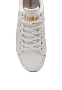 Vita sneakers tillverkade av slät syntetiskt material, med platta snören och en gyllene "Gola"-logotyp på tungan. Rundad tå och gummisulor.