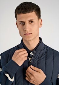 Marinblå vadderad jacka med ribbade ärmslut och ståkrage, med dragkedja och ett lager skjorta under med randigt mönster.